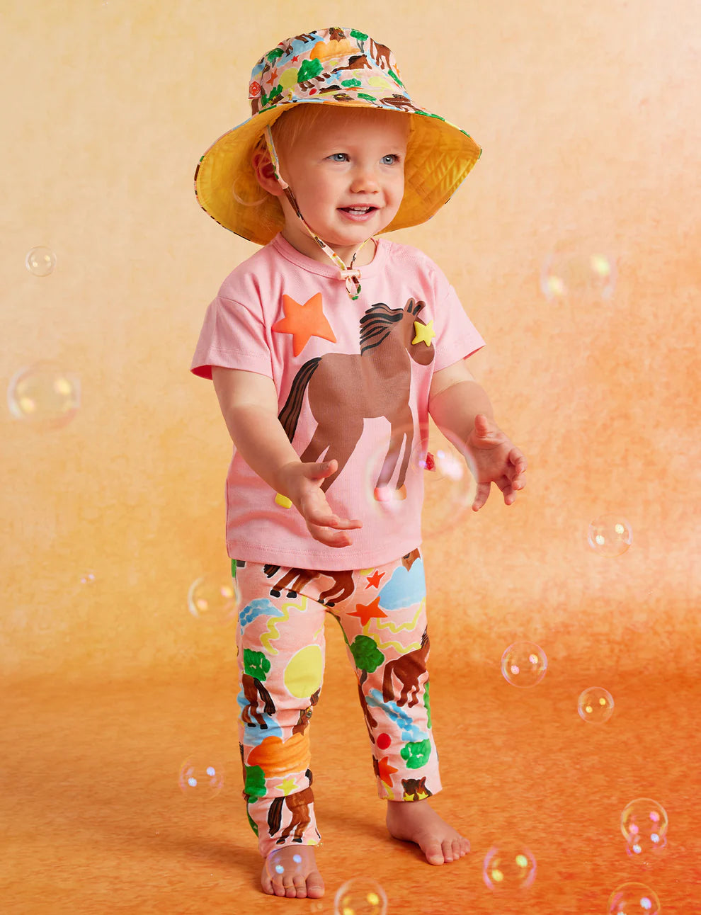 Child wearing a colorful hat and animal-themed shirt and leggings on a yellow background with bubbles.
