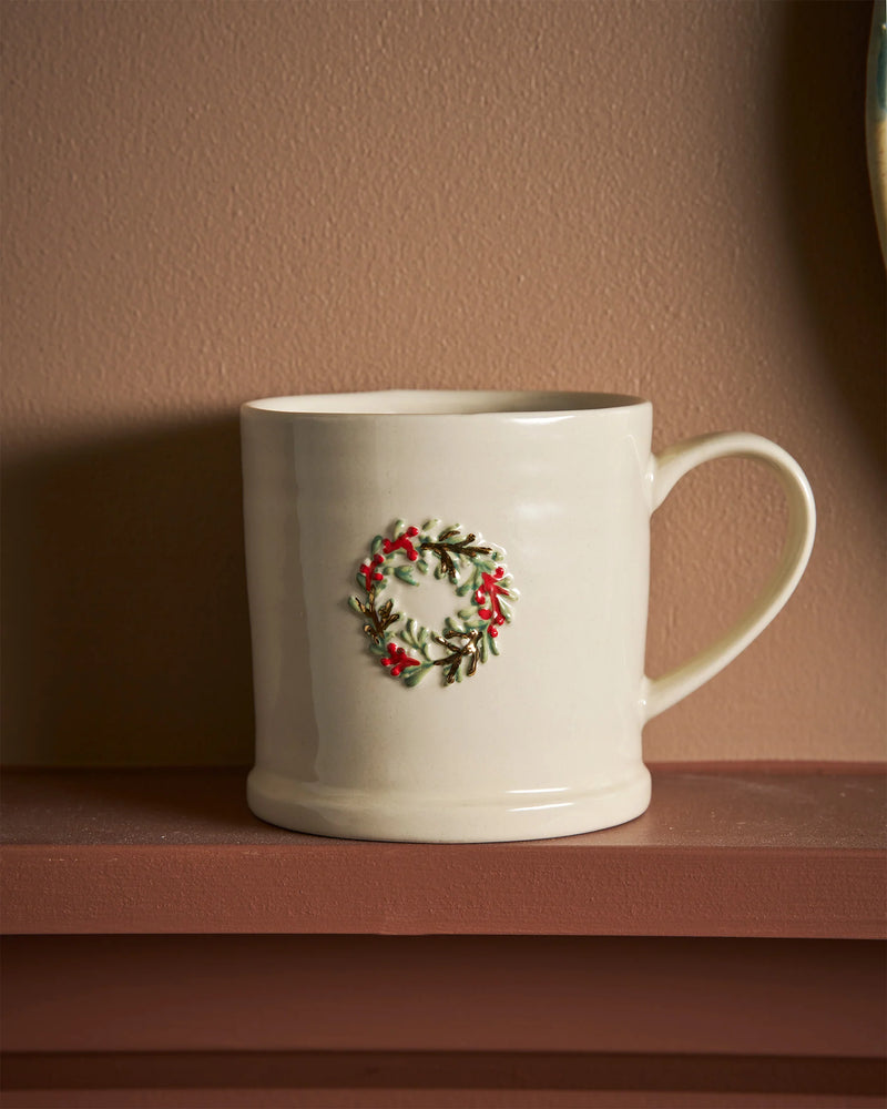 An off-white ceramic mug with a small green and red holly wreath design on the front