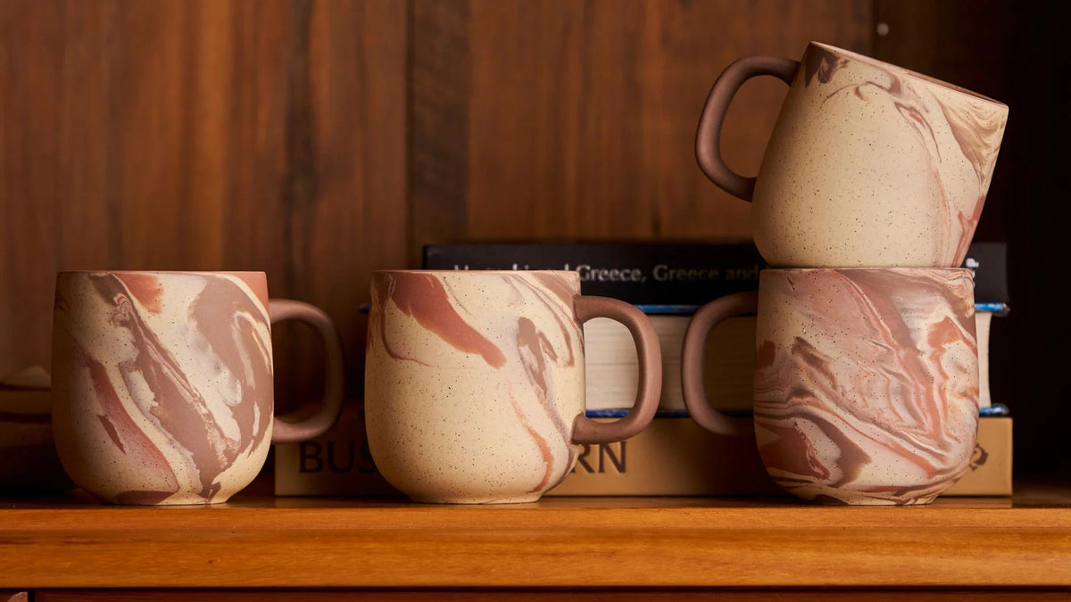 Four Robert Gordon Pottery ceramic mugs with a pink and beige marble pattern are displayed on a wooden shelf in front of a dark wood-paneled background.