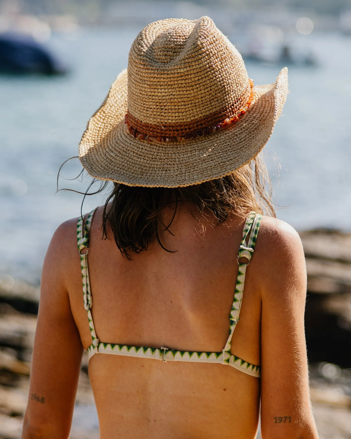 A woman viewed from behind wearing a wide-brimmed straw hat with a brown band and a green-and-white patterned bikini top.