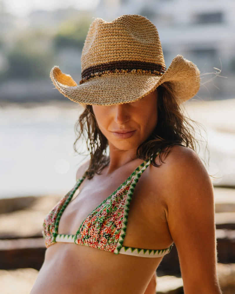 A woman wearing a woven straw cowboy hat with a beaded hat band and a patterned bikini top, photographed outdoors.