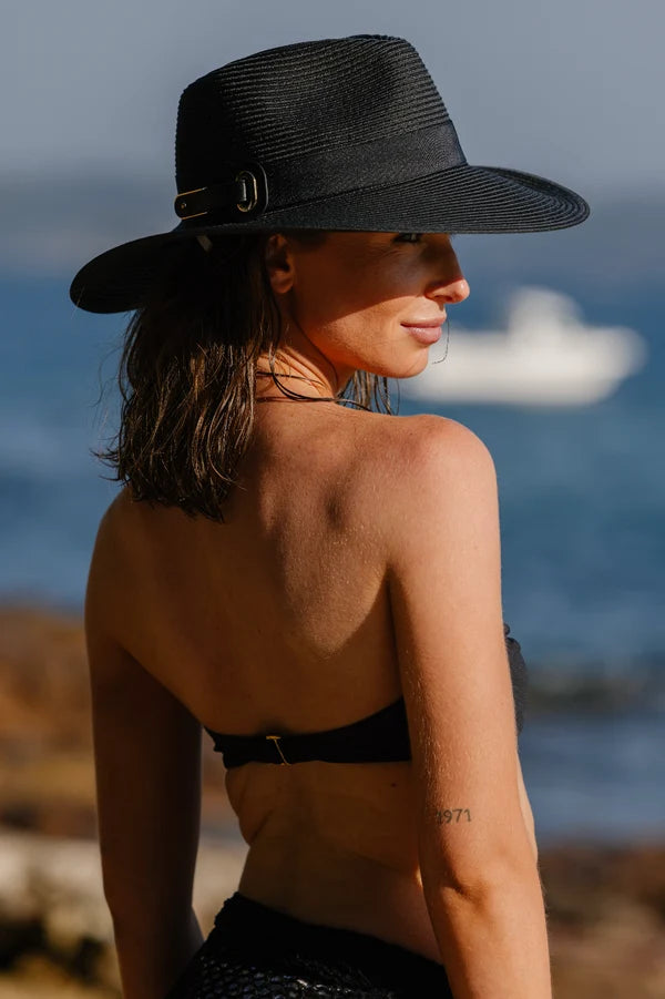 A woman wearing a wide-brimmed black straw hat with a gold buckle, photographed outdoors by the sea with a boat in the background.