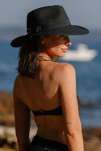 A woman wearing a wide-brimmed black straw hat with a gold buckle, photographed outdoors by the sea with a boat in the background.