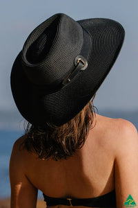 A woman viewed from behind wearing a black woven wide-brim sun hat with a small silver buckle detail.