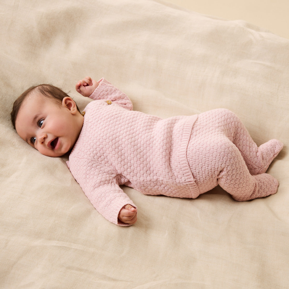 Baby in a pink textured onesie lying on a beige blanket