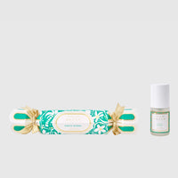 Decorative cracker with gold bow and green label next to a small bottle on a light gray background