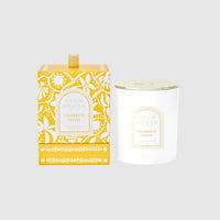 White candle with gold lid and decorative yellow packaging on a light gray background